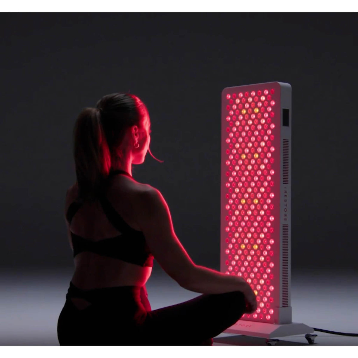 Woman seated in front of the iRESTORE Apex Pro 1500 full body red light panel on a floor stand with all LEDs lit red