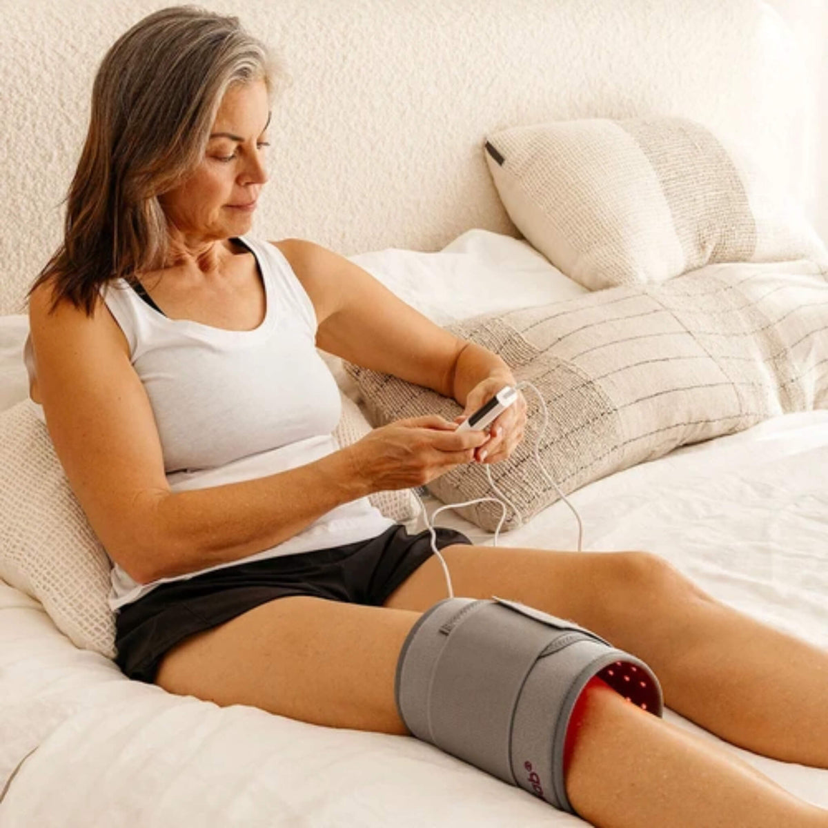 Woman on a bed with the NovaaLab Novaa Light Pad strapped around her knee, adjusting the remote control
