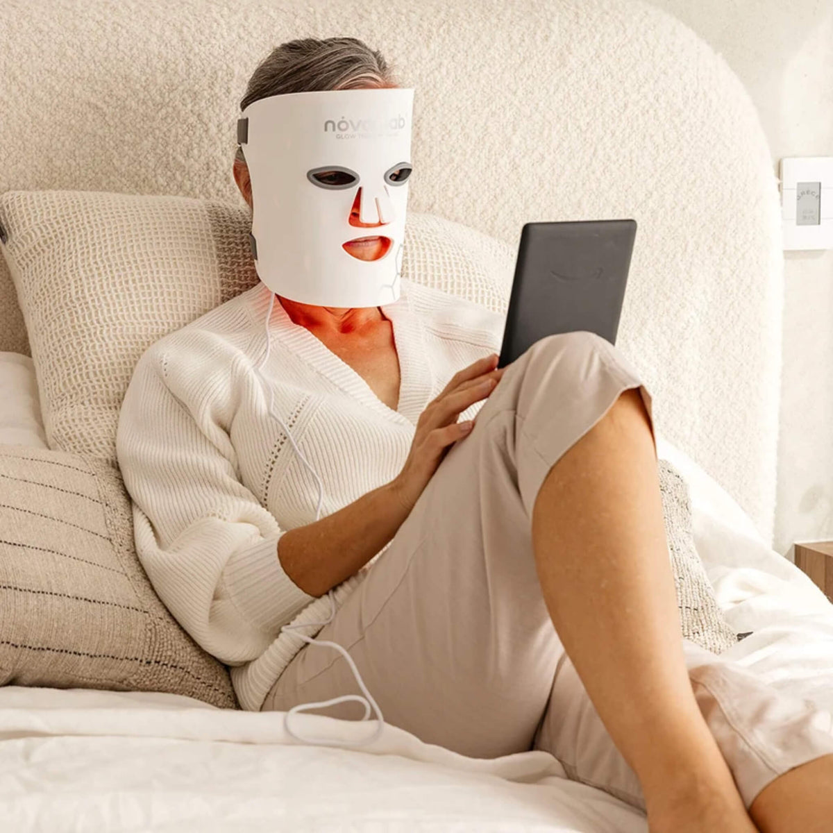 Woman relaxing in bed reading a tablet while wearing the NovaaLab Novaa Glow Therapy Mask