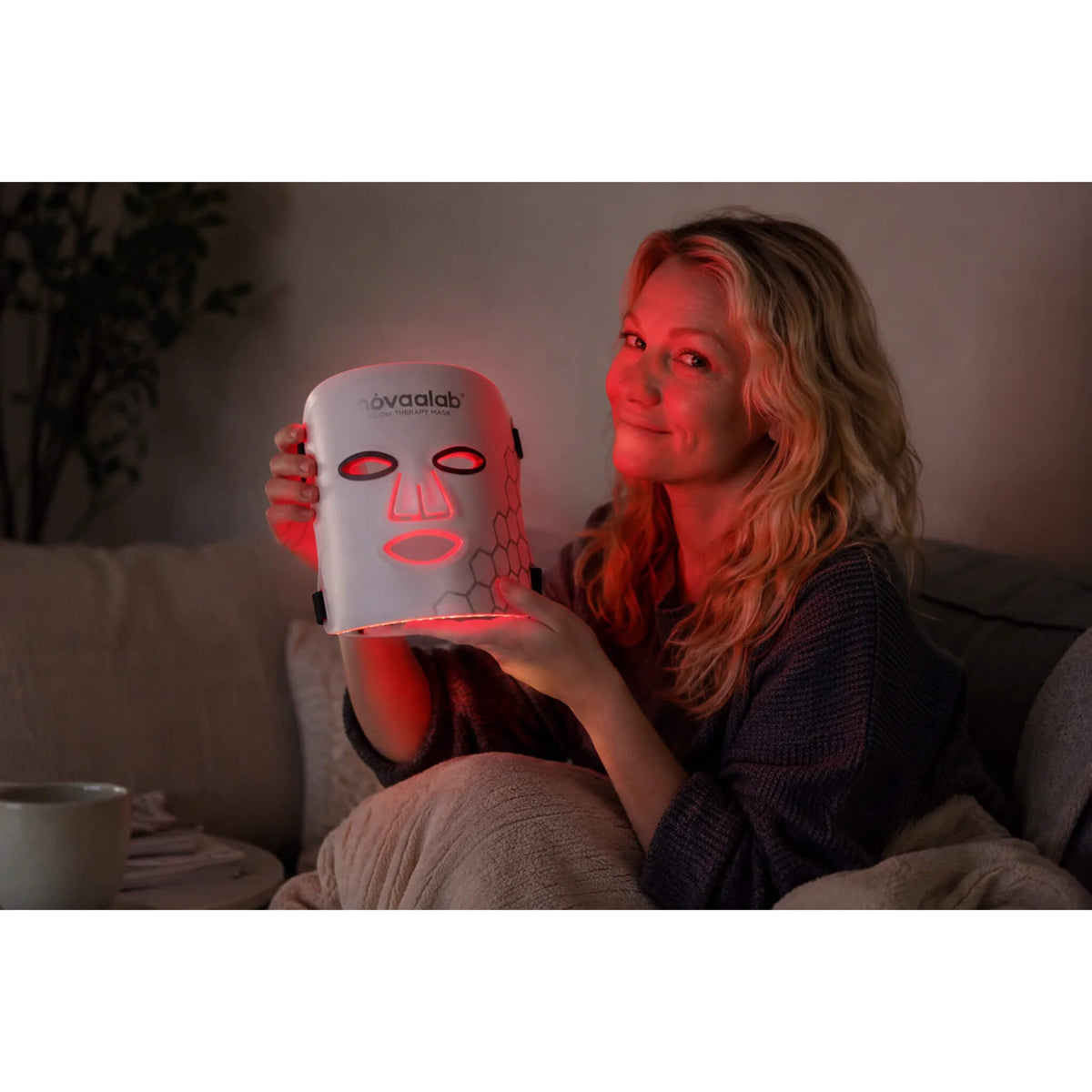 Smiling woman on the couch holding the NovaaLab Novaa Glow Therapy Mask in red light mode