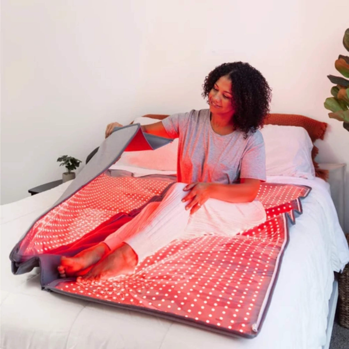 Woman sitting in the open NovaaLab Novaa Recovery Pod on a bed, lifting the upper half with the red LEDs on