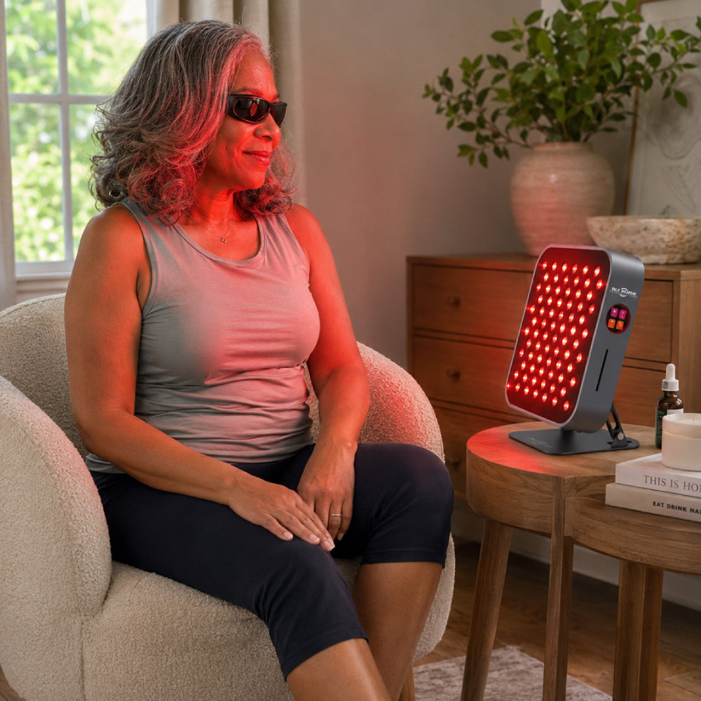Woman wearing eye protection seated next to the RLT Home Total Spectrum MINI Red Light Therapy Device on a side table at home